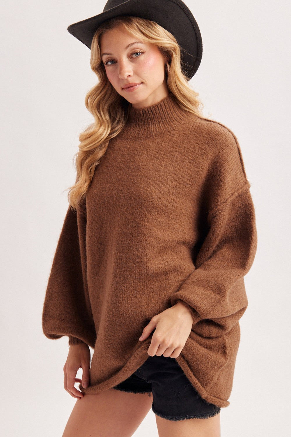woman wearing a brown oversized sweater with a high neckline and wide sleeves, paired with black shorts and a black hat