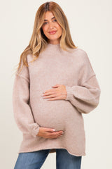 woman wearing a beige oversized maternity sweater with a high neck and holding her baby bump