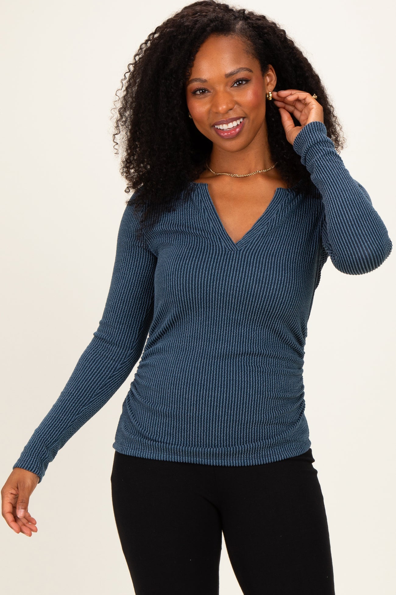 woman wearing a navy blue long sleeve fitted top with a v-neck and ribbed texture, smiling and posing with one hand in her hair