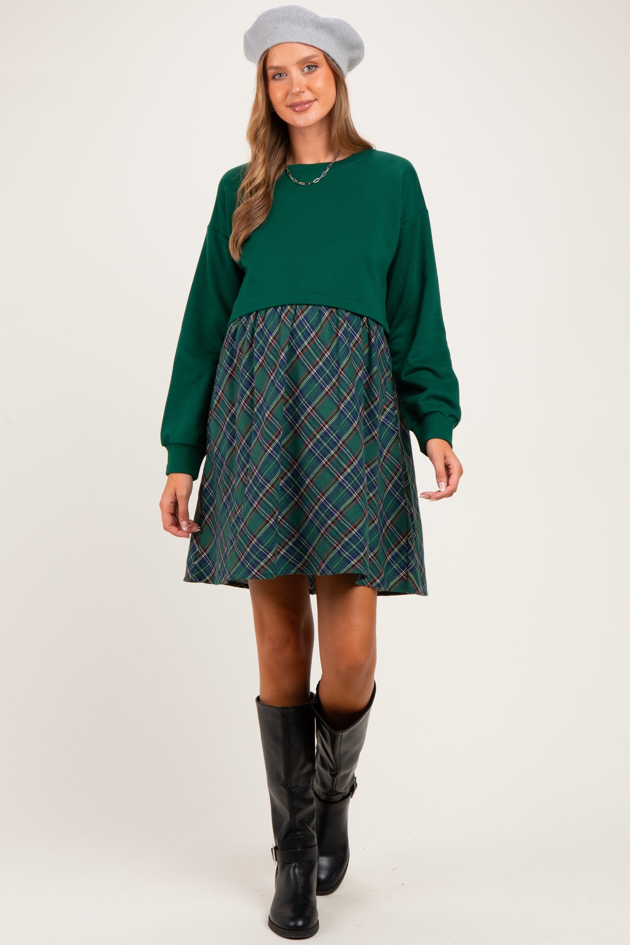 woman wearing a green maternity dress with long sleeves and a plaid skirt, styled with a gray beret and black knee-high boots