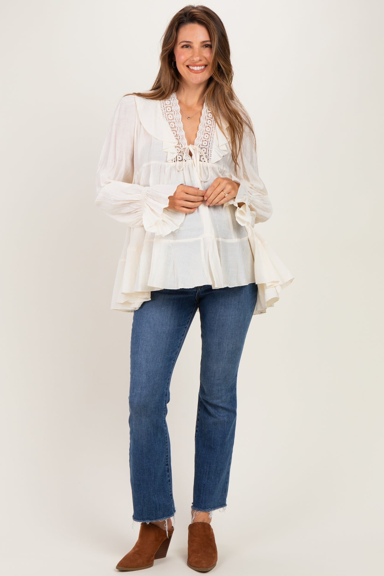woman wearing a cream maternity blouse with lace details and flared sleeves, paired with dark blue jeans and brown ankle boots