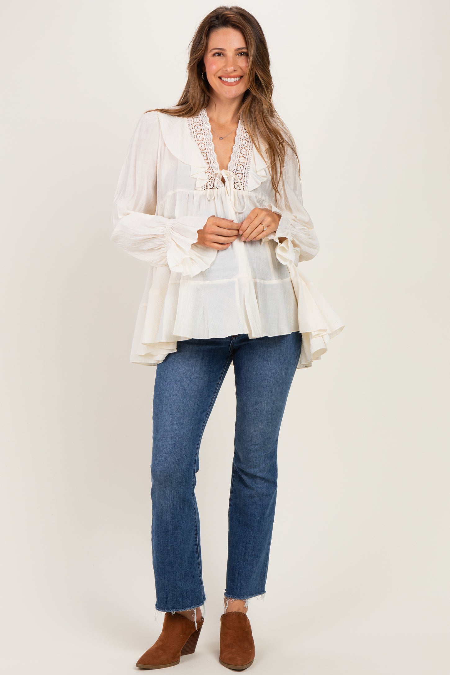 woman wearing a cream maternity blouse with lace details and flared sleeves, paired with dark blue jeans and brown ankle boots