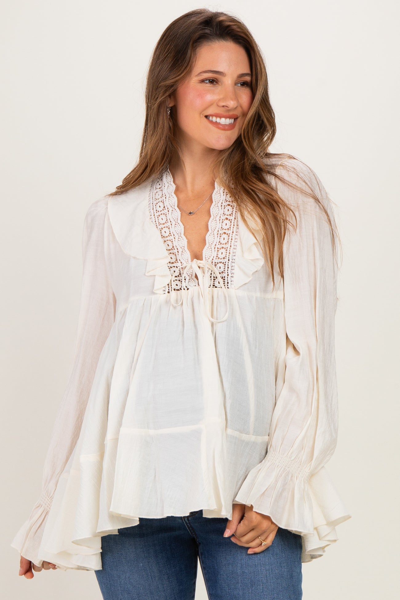woman wearing a cream maternity blouse with ruffle details and a lace-up neckline, styled with blue jeans