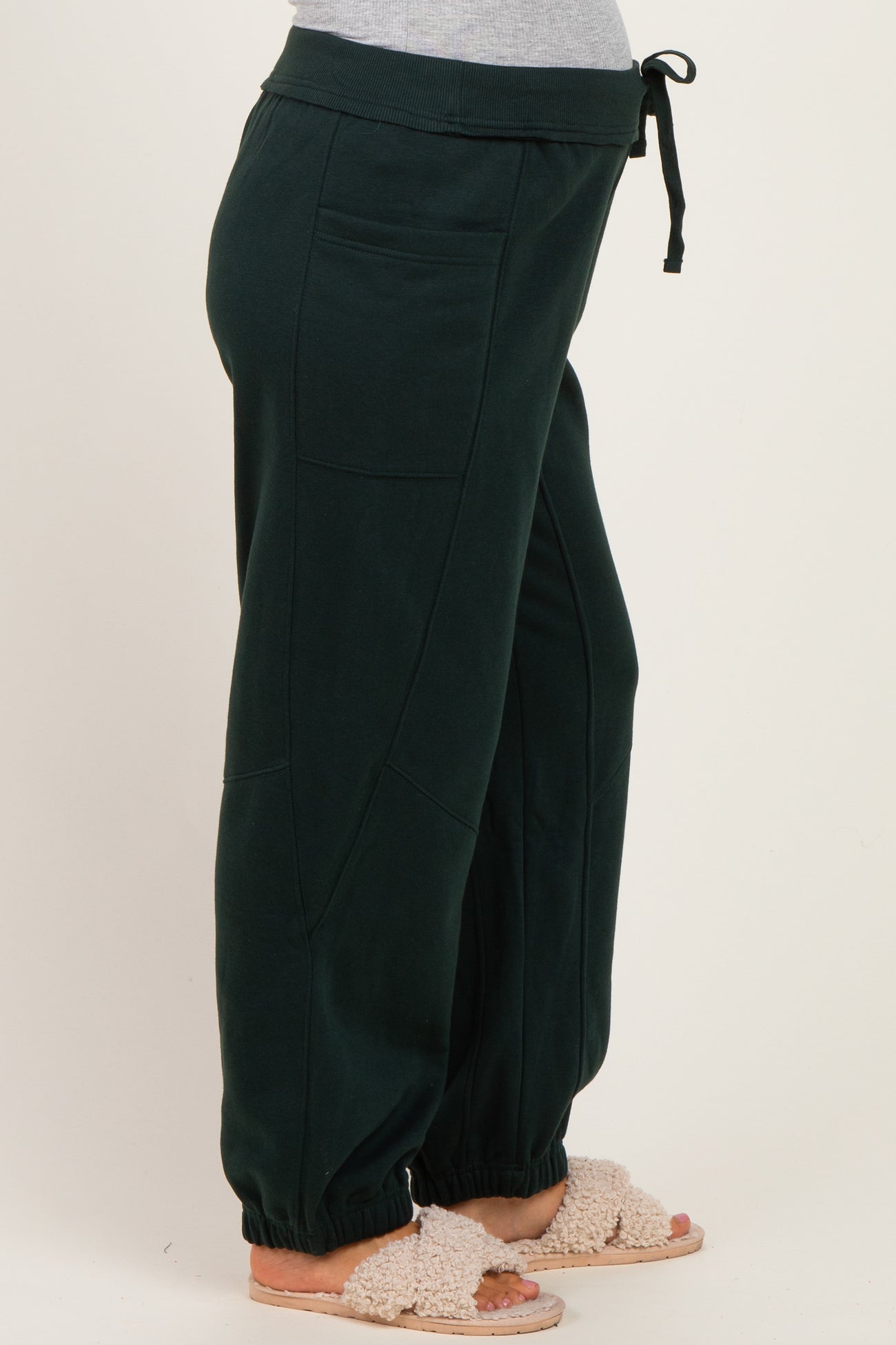 woman wearing dark green maternity sweatpants with side pockets and elastic cuffs, styled with a casual top and slippers