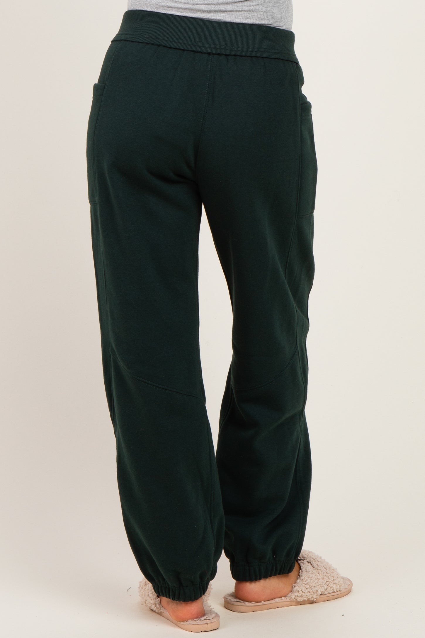 woman wearing dark green maternity jogger pants with side pockets and elastic cuffs, standing with back facing camera