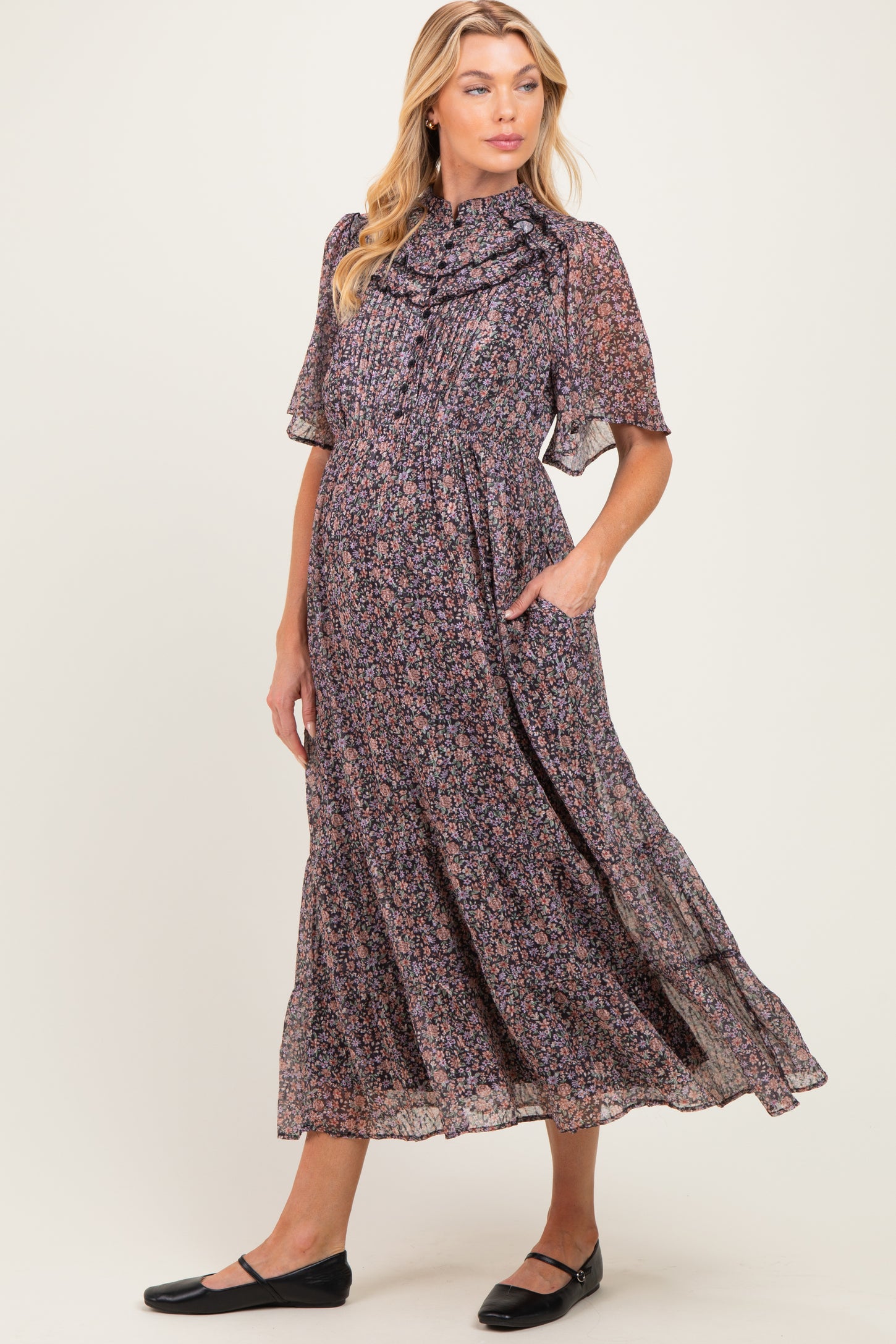 woman wearing a floral print maternity dress with short sleeves and a ruffled neckline, featuring pockets and a flowing skirt