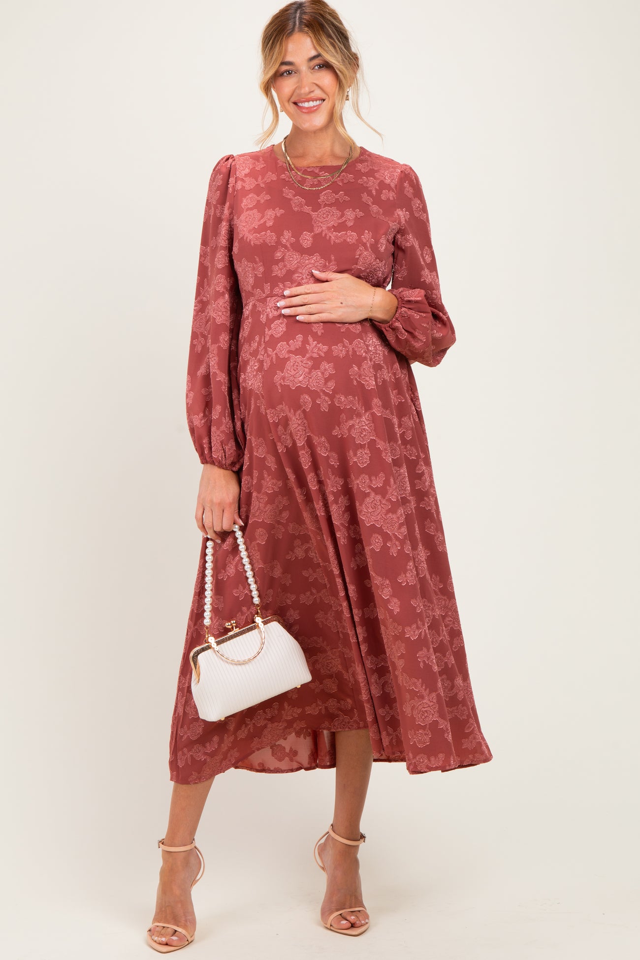 woman wearing a rust floral long sleeve maternity dress and holding her baby bump with a white handbag