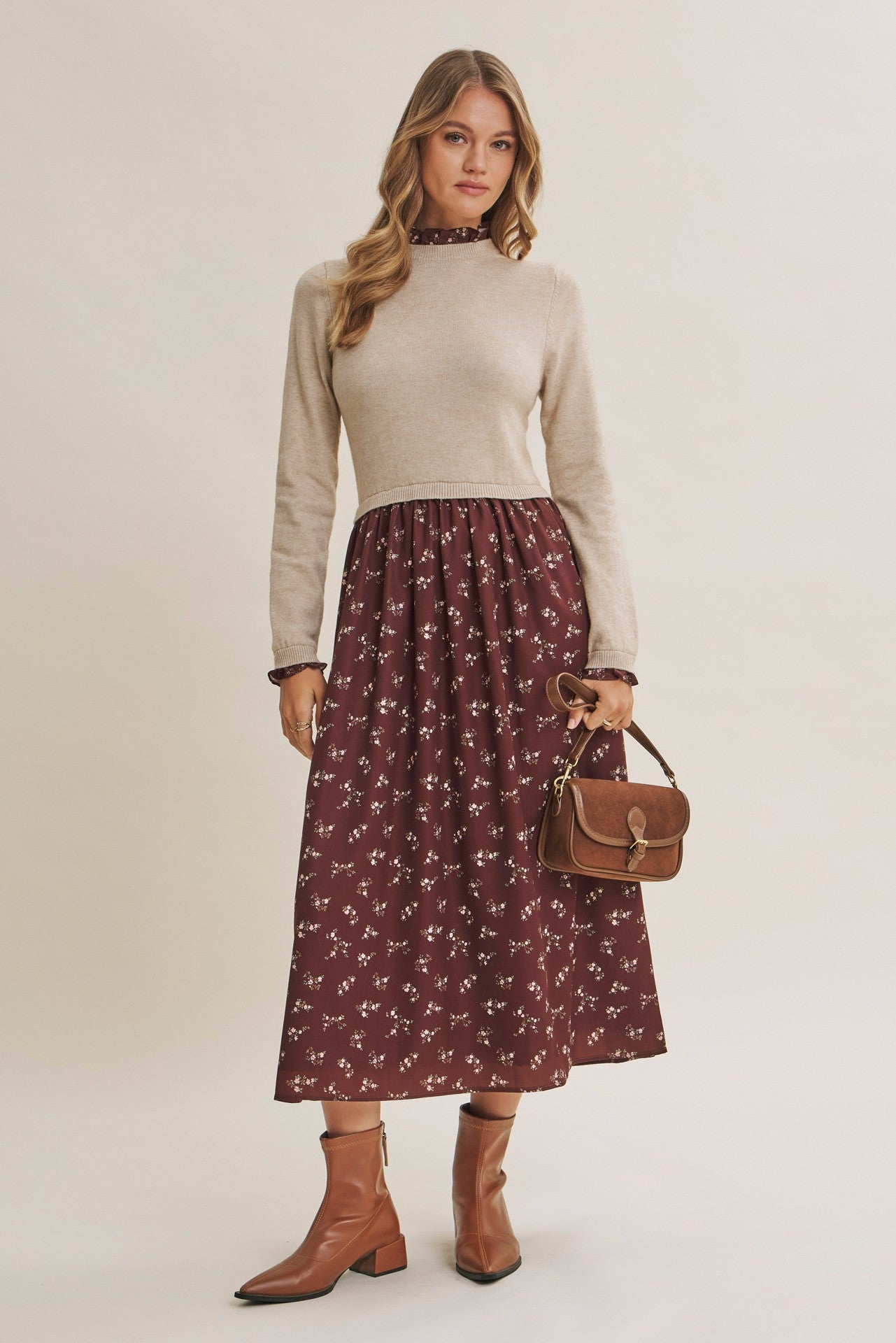 woman wearing a beige long sleeve knit top and a burgundy floral midi skirt, holding a brown handbag