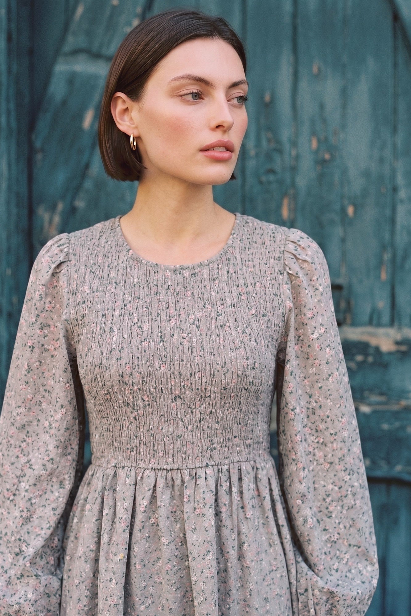 woman wearing a smocked floral print dress with long sleeves and a round neckline standing against a teal wooden background