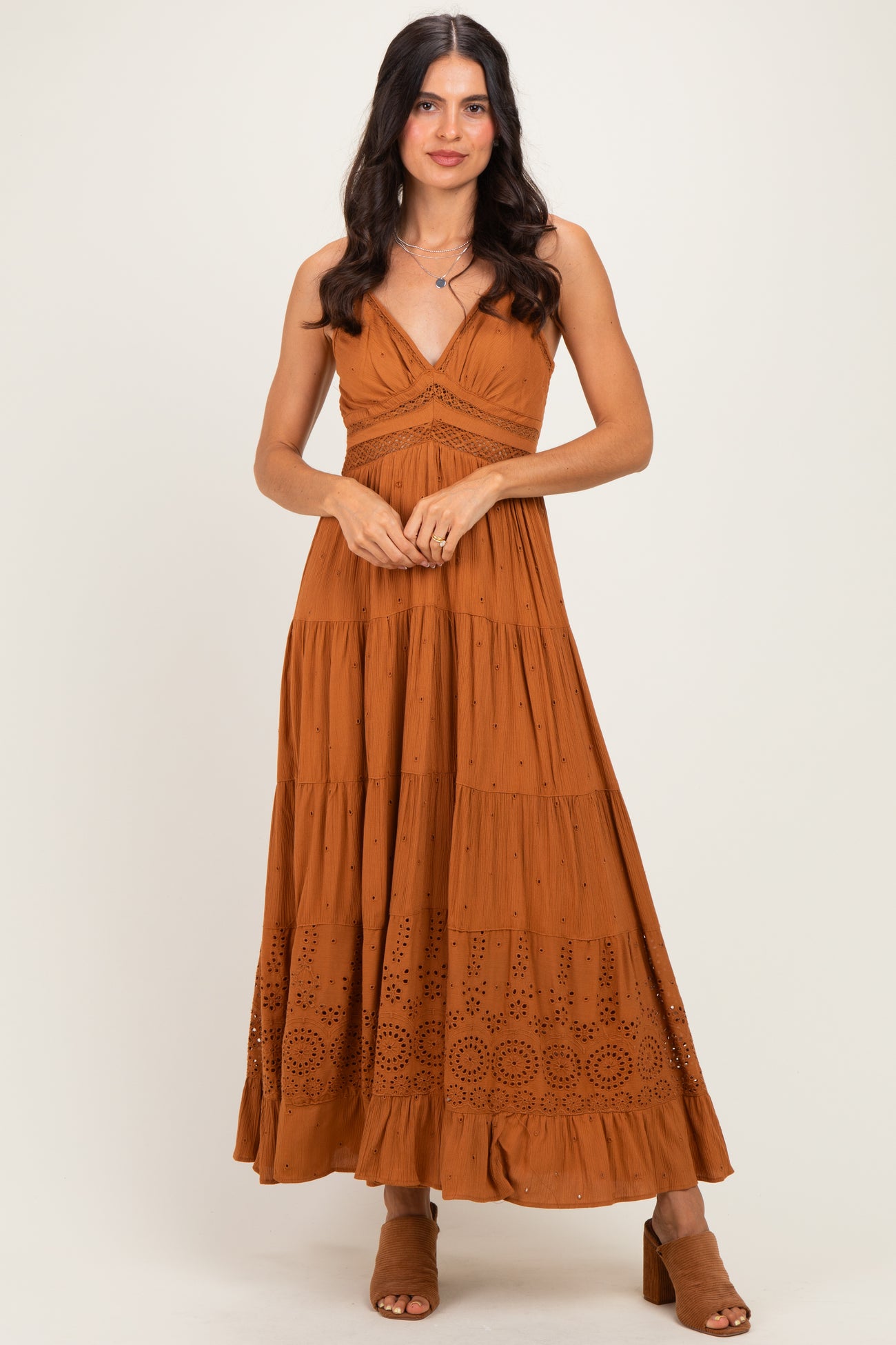 woman wearing a rust-colored maxi dress with a tiered design and eyelet details, standing with a slight pose