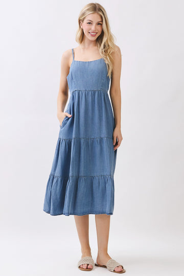 woman wearing a sleeveless denim midi dress with tiered ruffles and pockets, standing and smiling