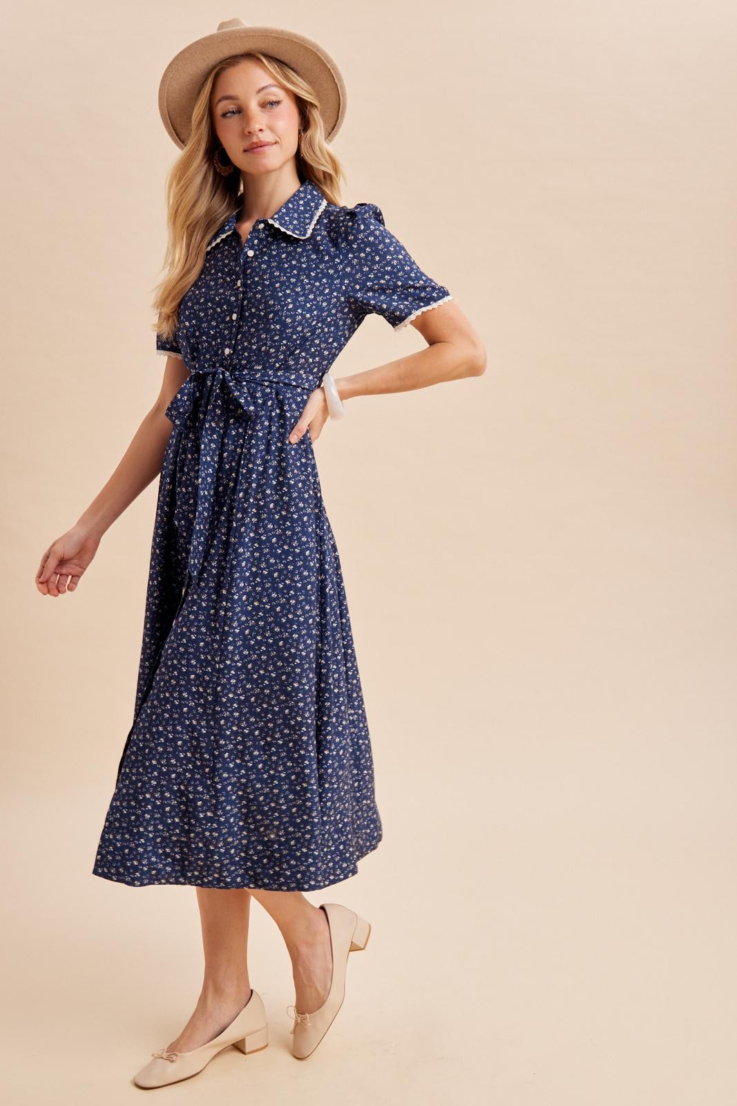 woman wearing a navy blue floral print midi dress with short sleeves and a waist tie, styled with a beige hat and flats