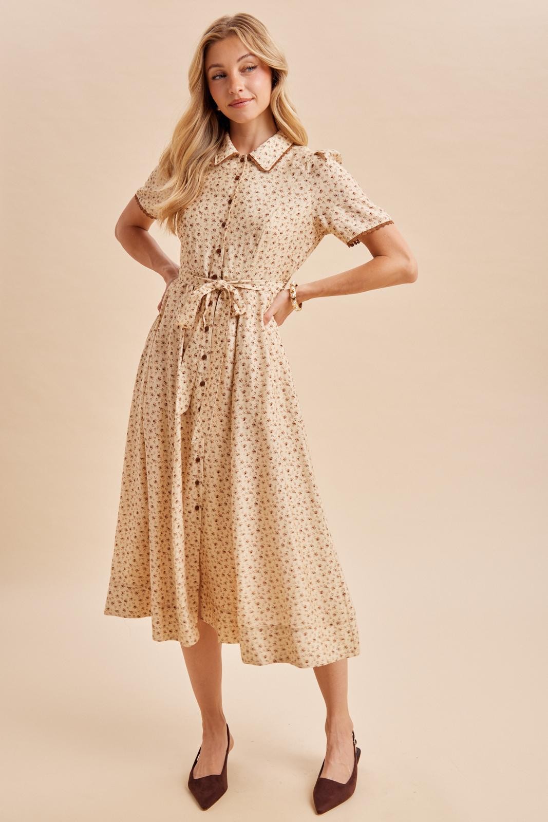woman wearing a beige floral midi dress with short sleeves and a tie waist detail, posing with one hand on her hip