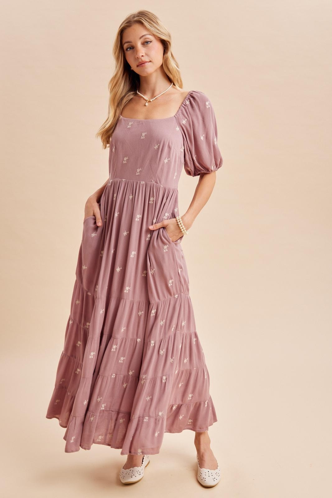 woman wearing a mauve puff sleeve maxi dress with floral pattern and pockets, standing against a neutral background