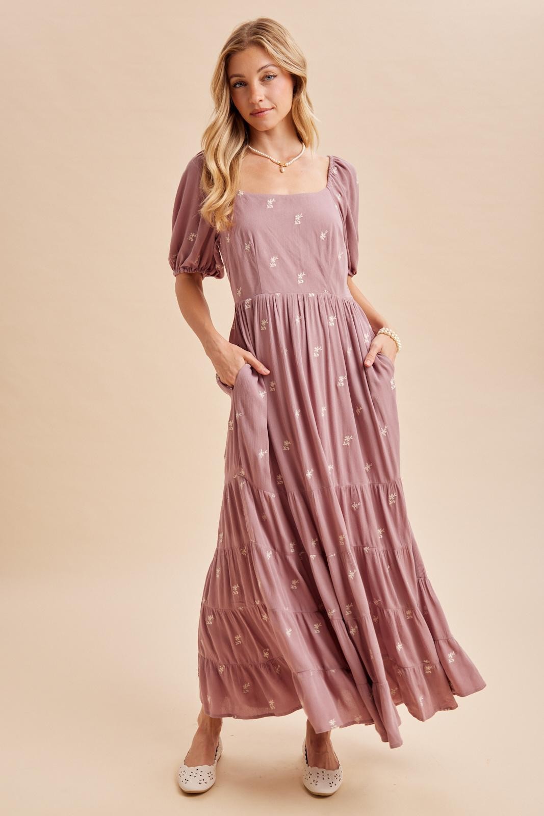 woman wearing a mauve maxi dress with puff sleeves and floral embroidery, featuring pockets and a flowing skirt