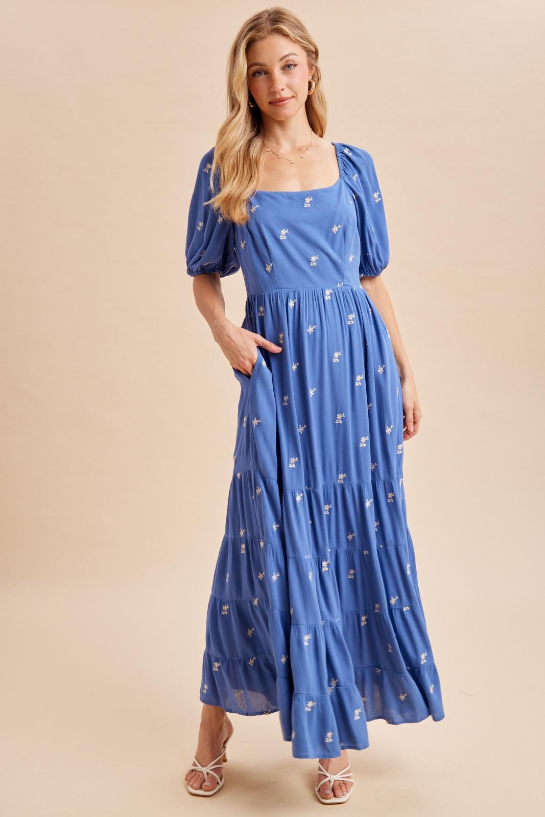 woman wearing a blue floral print maxi dress with puff sleeves and pockets, standing against a neutral background