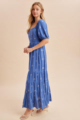 woman wearing a blue maxi dress with puff sleeves and floral embroidery, standing in profile against a neutral backdrop
