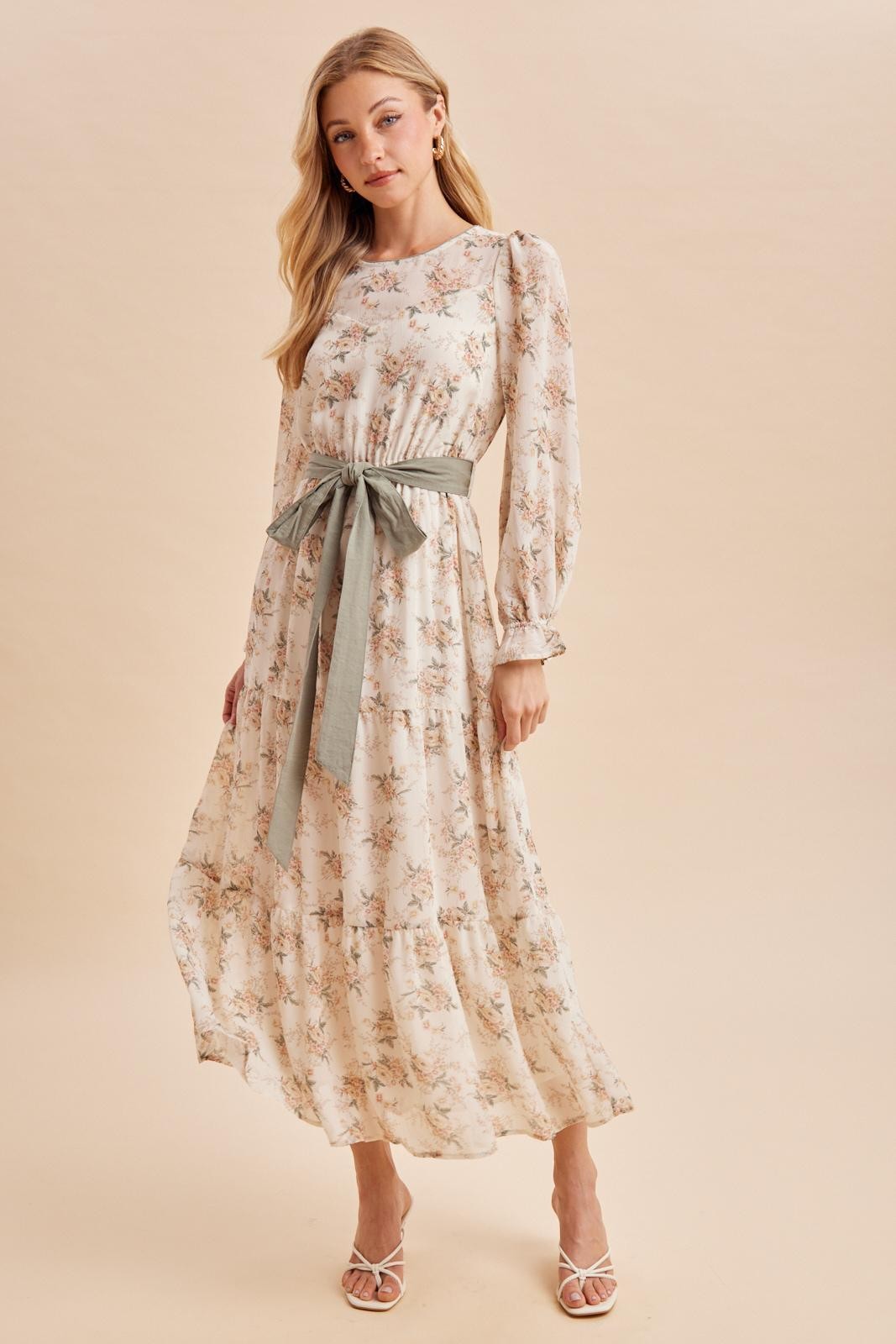woman wearing a floral maxi dress with long sleeves and a tie waist in soft pastel colors, posing against a neutral background