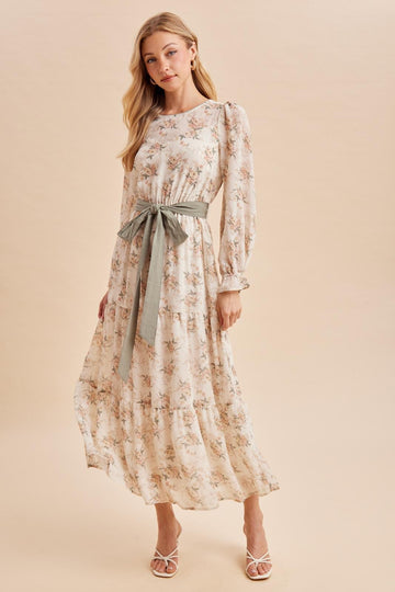 woman wearing a floral maxi dress with long sleeves and a tie waist in soft pastel colors, posing against a neutral background