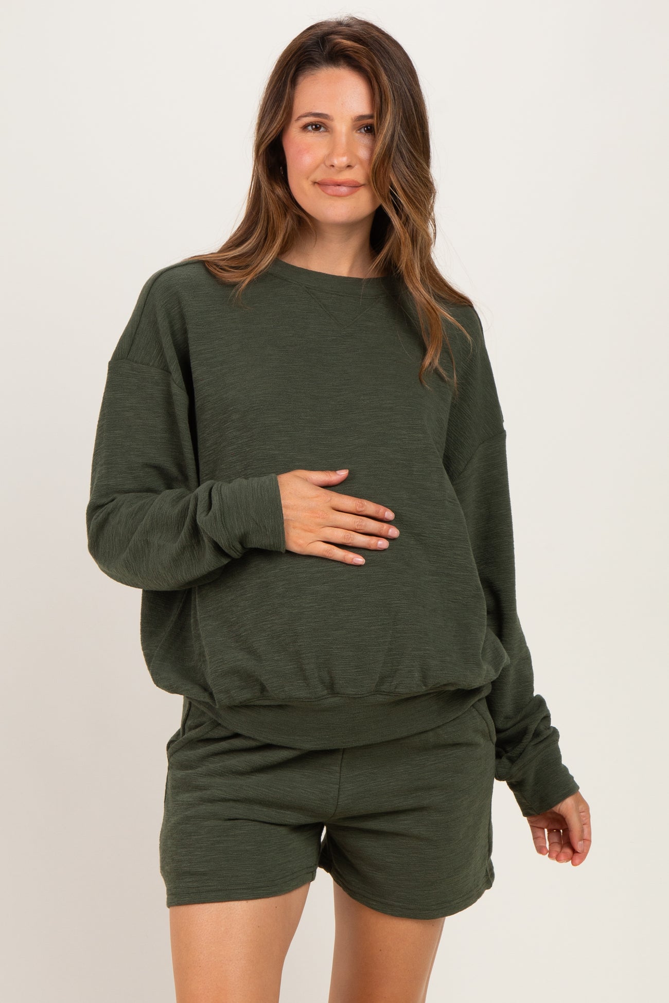 woman wearing a dark green maternity sweatshirt and matching shorts, holding her baby bump