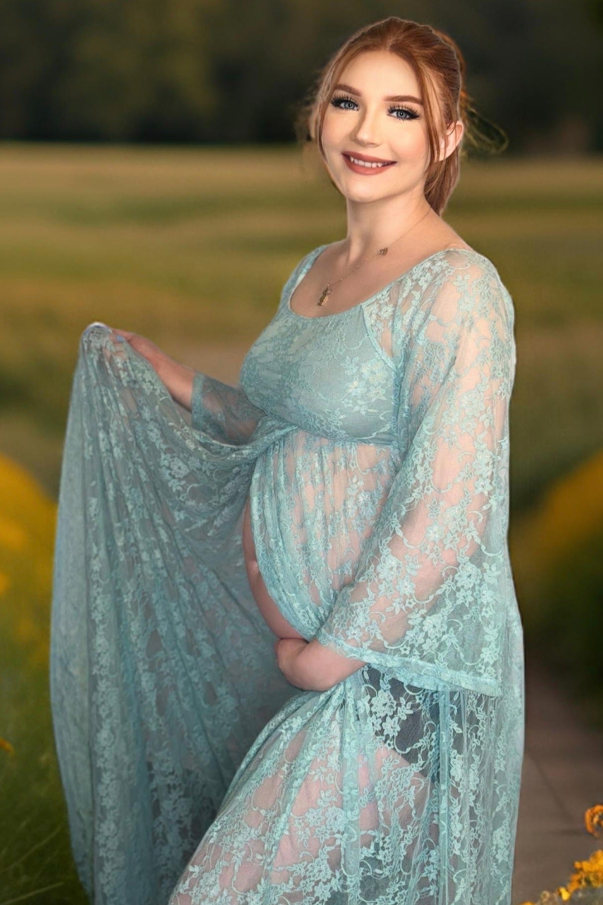 woman wearing a light blue lace maternity gown and holding her baby bump in a field setting