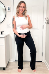 woman wearing a white maternity tank top and black maternity pants, showcasing her baby bump while smiling