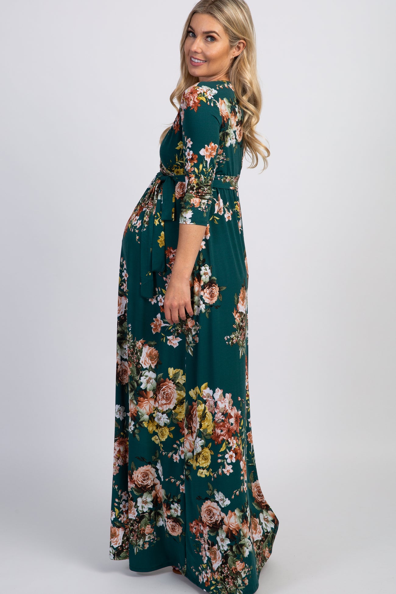 woman wearing a teal floral maternity maxi dress with three-quarter sleeves and a tied waist, showing her baby bump