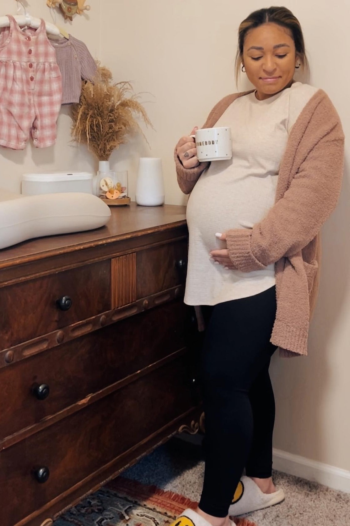 woman wearing a light beige maternity top, black leggings, and a cozy cardigan, holding a mug and showing her baby bump.
