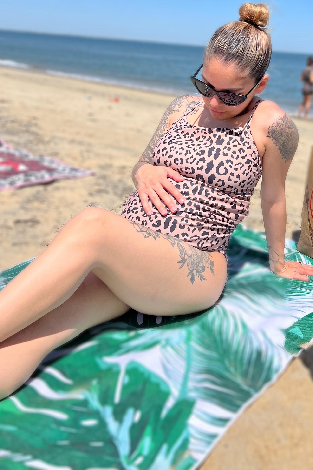 woman wearing a leopard print maternity swimsuit, sitting on a beach towel and holding her baby bump