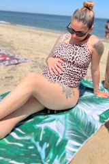 woman wearing a leopard print maternity swimsuit, sitting on a beach towel and holding her baby bump