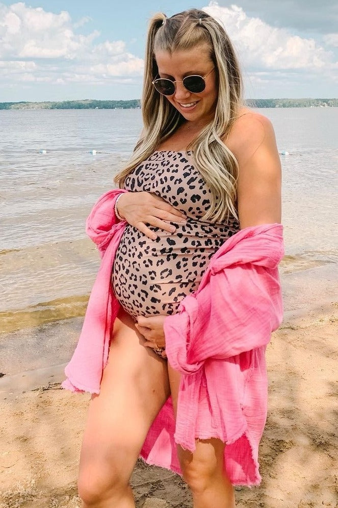 woman wearing a leopard print maternity swimsuit and a pink cover-up, holding her baby bump by the water