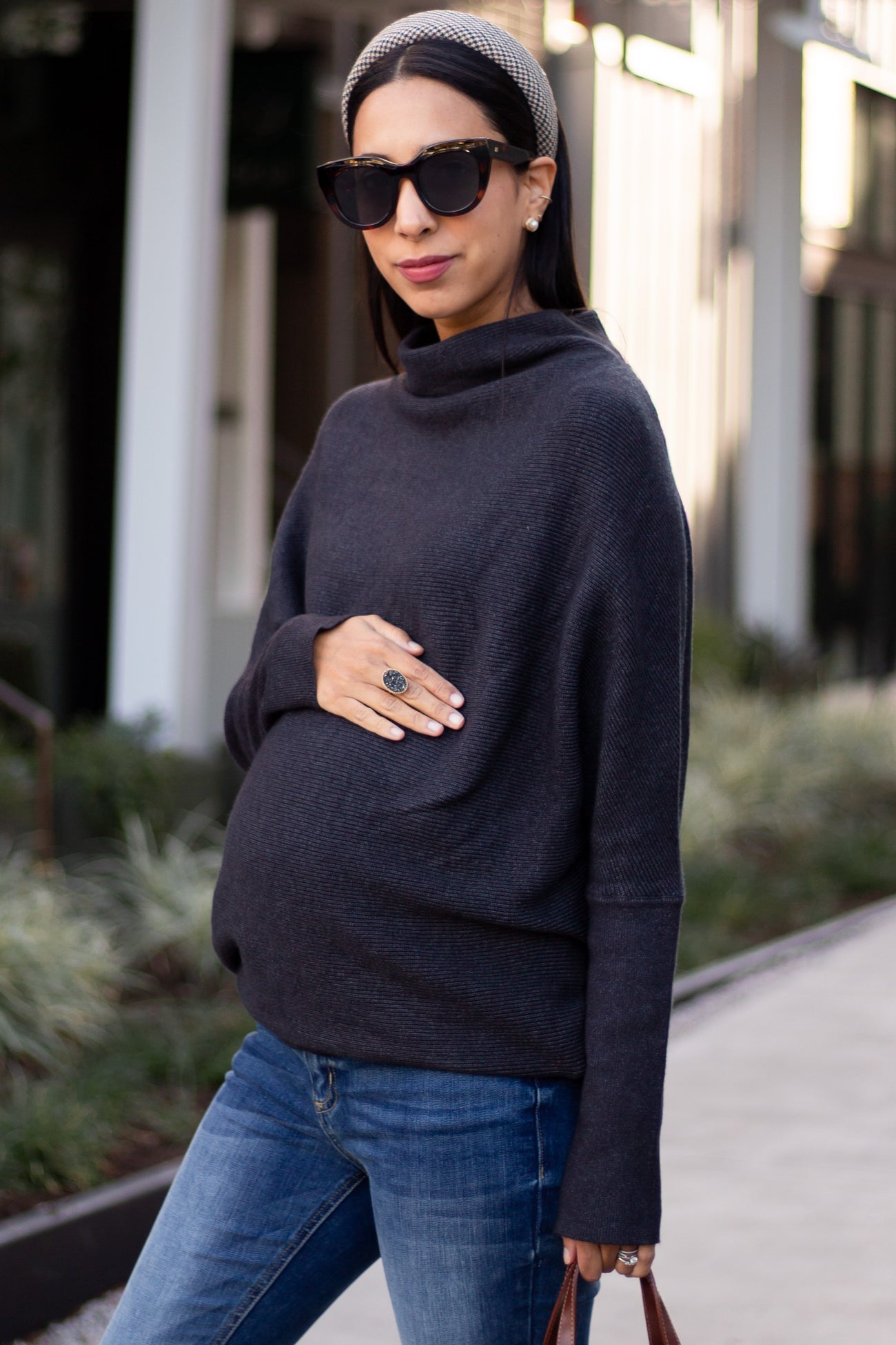 woman wearing a dark gray maternity turtleneck sweater and holding her baby bump outside in casual attire
