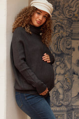 woman wearing a dark gray maternity turtleneck sweater and blue jeans, gently cradling her baby bump