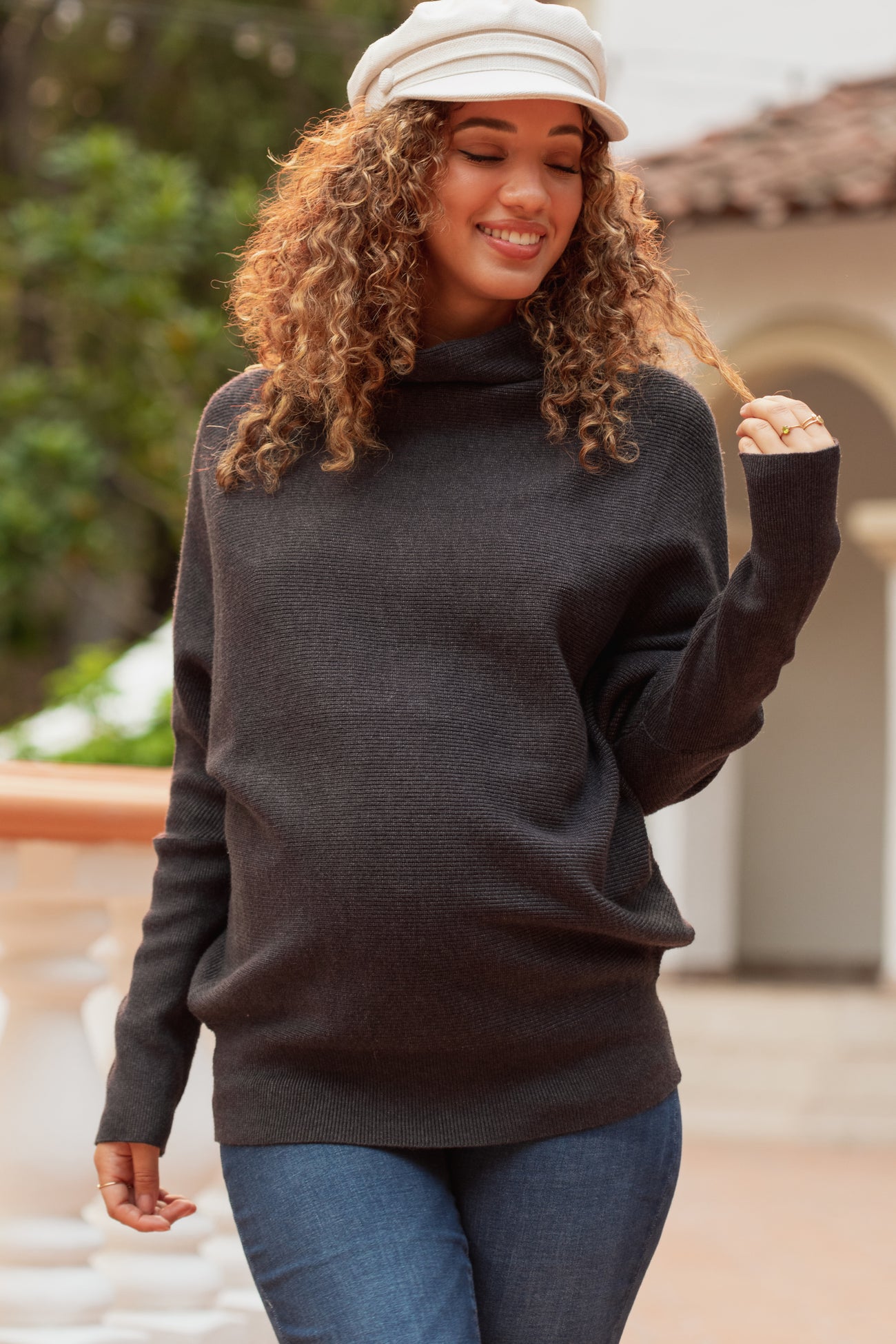 woman wearing a dark gray maternity sweater with a cowl neck and holding a strand of her curly hair, showing her baby bump
