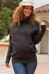 woman wearing a dark gray maternity sweater with a cowl neck and holding a strand of her curly hair, showing her baby bump