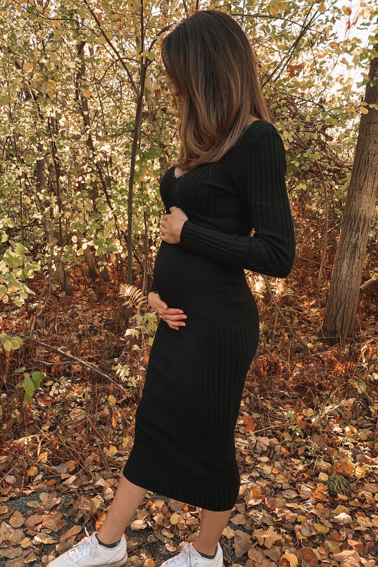 woman wearing a black ribbed maternity dress and holding her baby bump, standing in a forest with autumn leaves