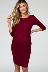 Pregnant woman wearing a burgundy three-quarter sleeve pleated maternity dress against a grey background