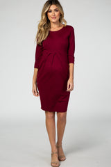 woman wearing a burgundy maternity dress with three-quarter sleeves and a pleated front detail, showing a baby bump