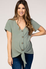 woman wearing a sage green maternity top with short sleeves, button details, and a front tie, showcasing her baby bump