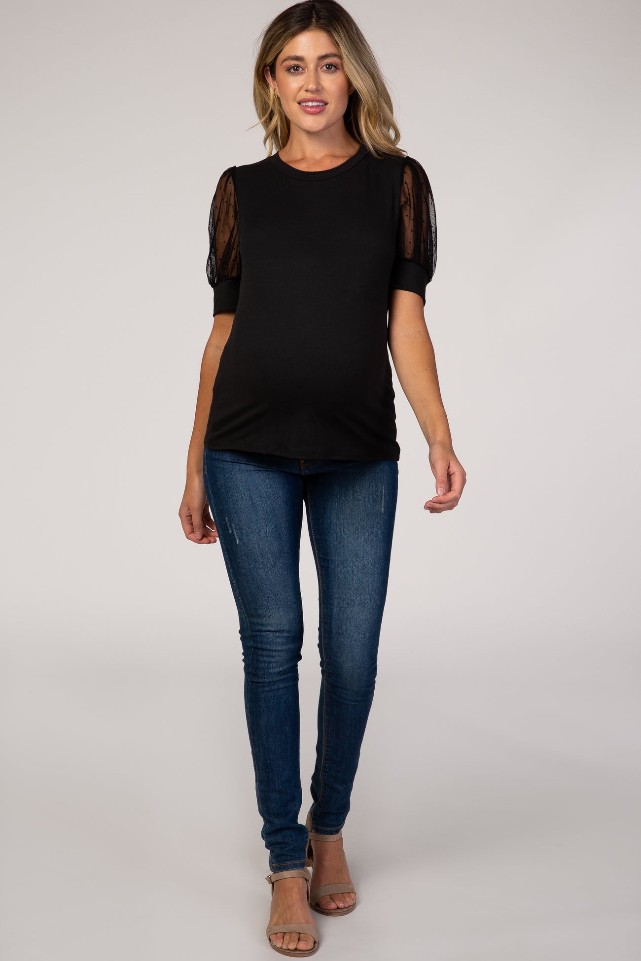 woman wearing a black maternity top with sheer puff sleeves and a visible baby bump, paired with blue skinny jeans