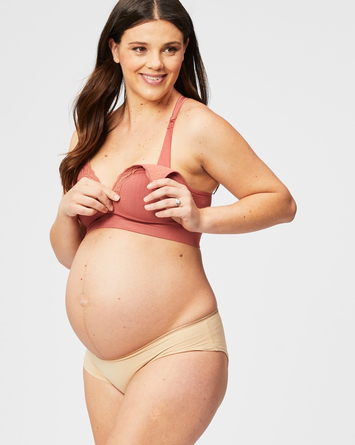 woman wearing a rust-colored maternity bra and nude maternity briefs, holding open the bra to show her baby bump