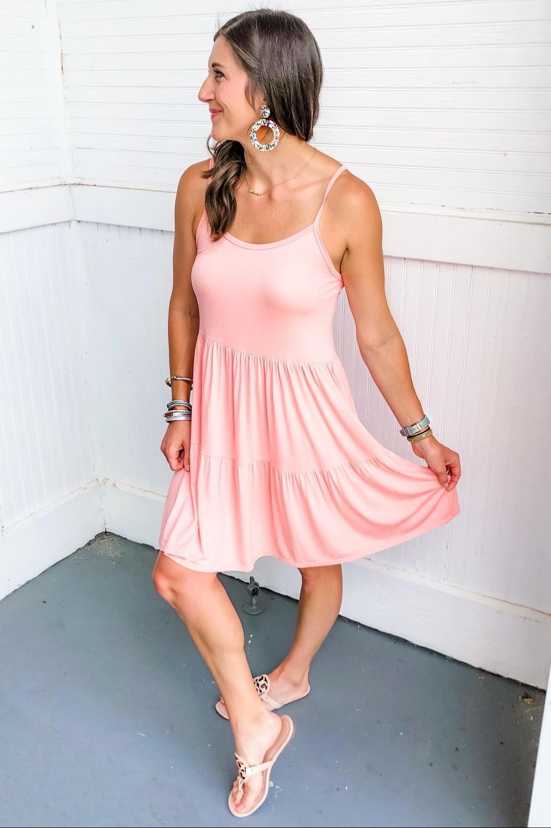 woman wearing a light pink spaghetti strap tiered dress and sandals, standing in a bright room with a smile.
