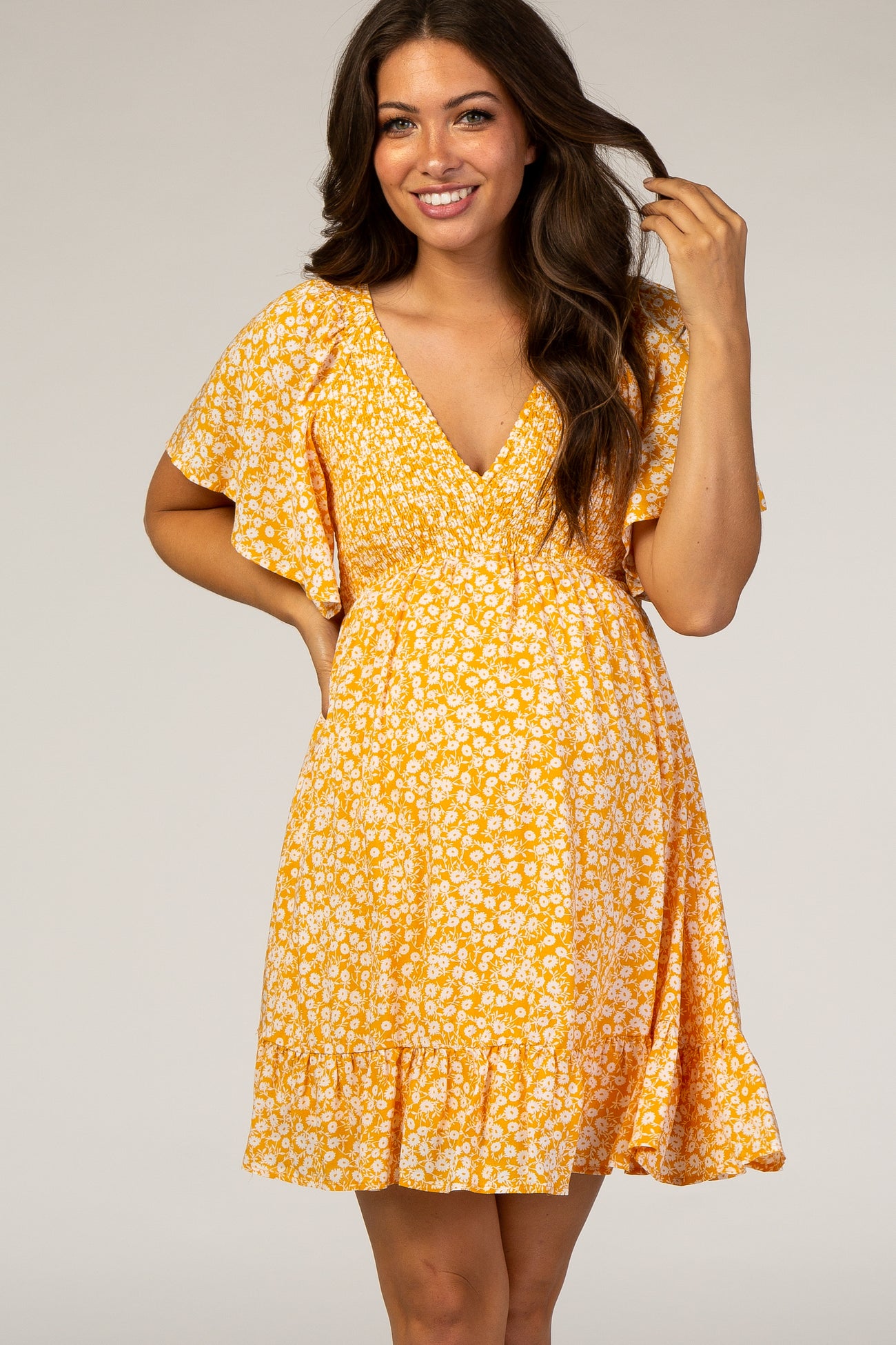 woman wearing a yellow floral maternity dress with a v-neck and flutter sleeves, smiling and holding her hair