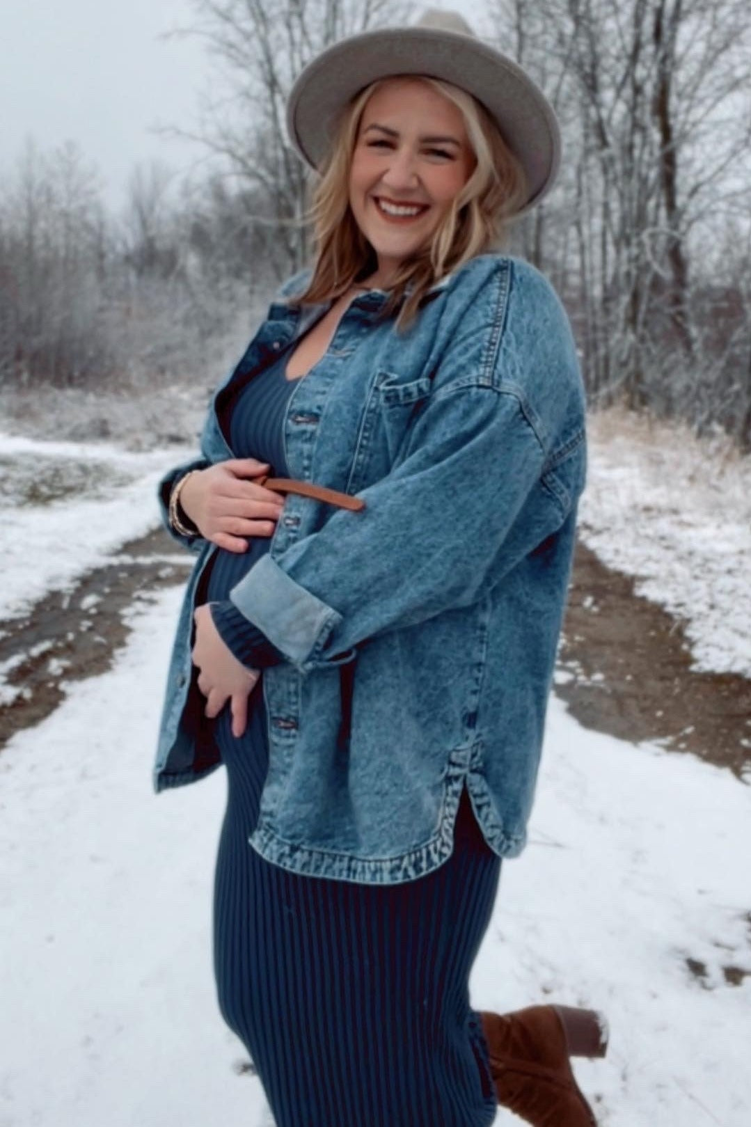 woman wearing a navy striped maternity dress and a denim jacket, holding her baby bump in a snowy outdoor setting