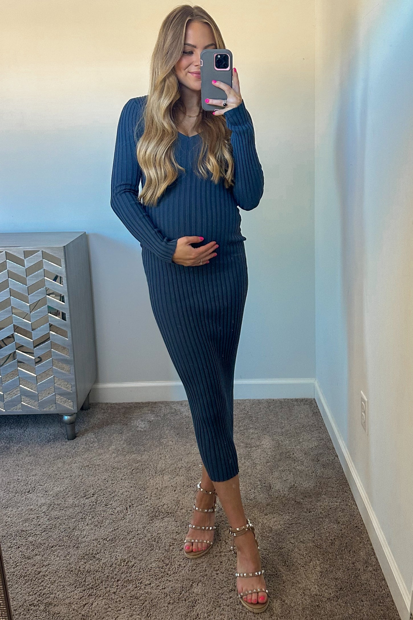woman wearing a dark blue ribbed maternity dress and holding her baby bump while standing in front of a mirror