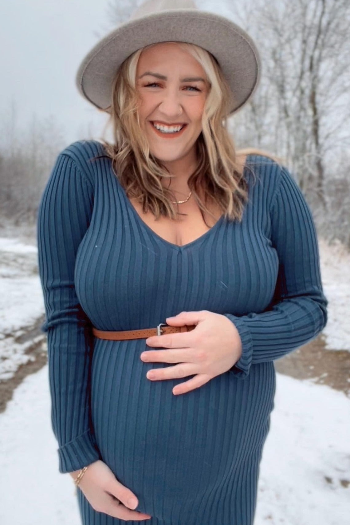 smiling pregnant woman in blue ribbed dress and wide brim hat holding her baby bump in a snowy outdoor setting