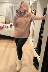 woman wearing a beige maternity sweater with a cowl neck and black leggings, posing in a mirror with a baby bump visible