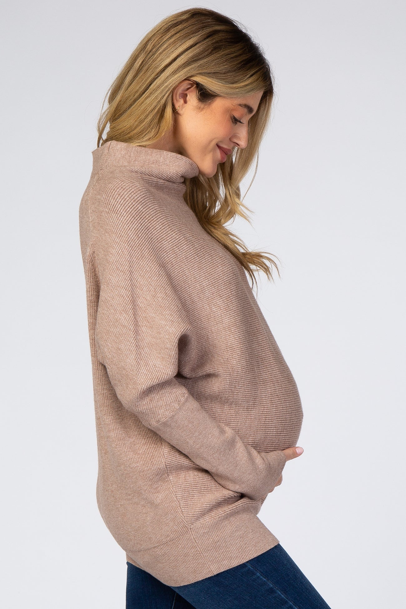 woman wearing a light brown oversized turtleneck maternity sweater and holding her baby bump