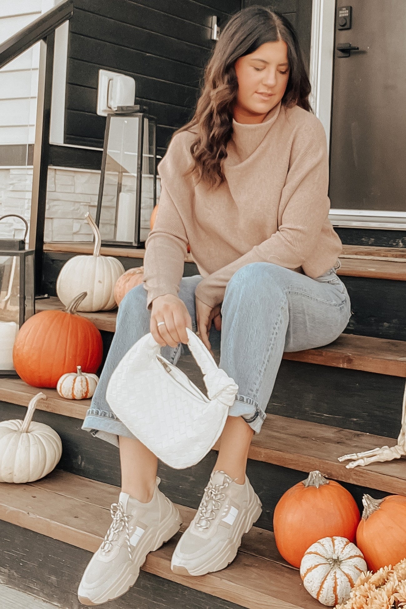 woman wearing a beige maternity sweater and light blue cuffed jeans, sitting on steps surrounded by pumpkins, holding a white bag