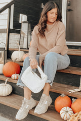 woman wearing a beige maternity sweater and light blue cuffed jeans, sitting on steps surrounded by pumpkins, holding a white bag